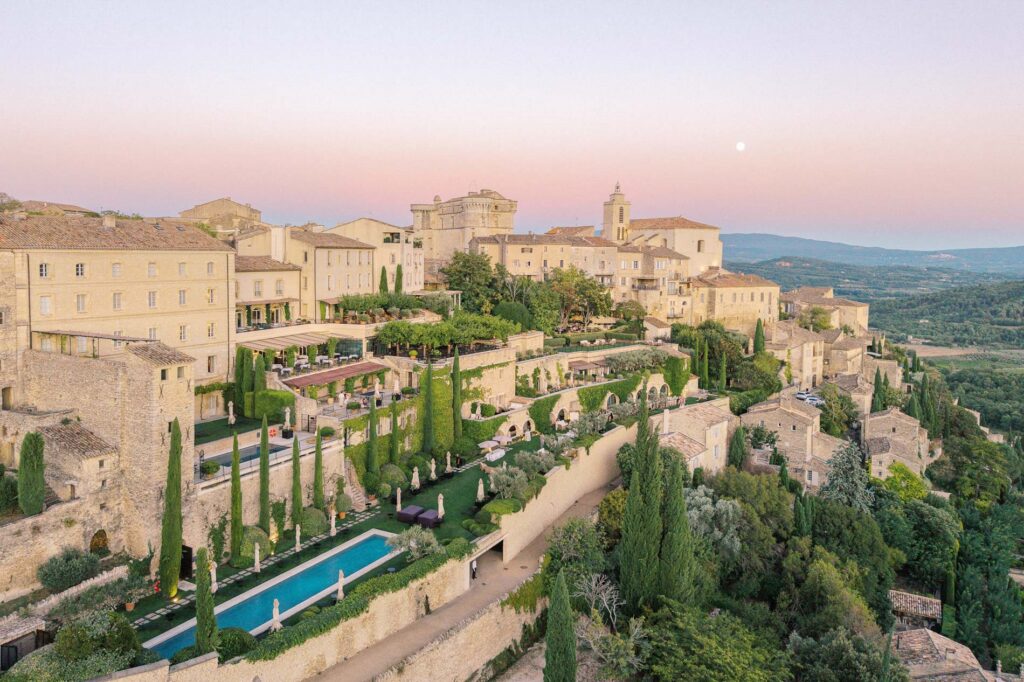 Aerial sunset view of Airelles Gordes, La Bastide overlooking the Luberon Valley, with terraced gardens, a long pool, cypress trees, and a quiet luxury wedding reception visible in the distance.