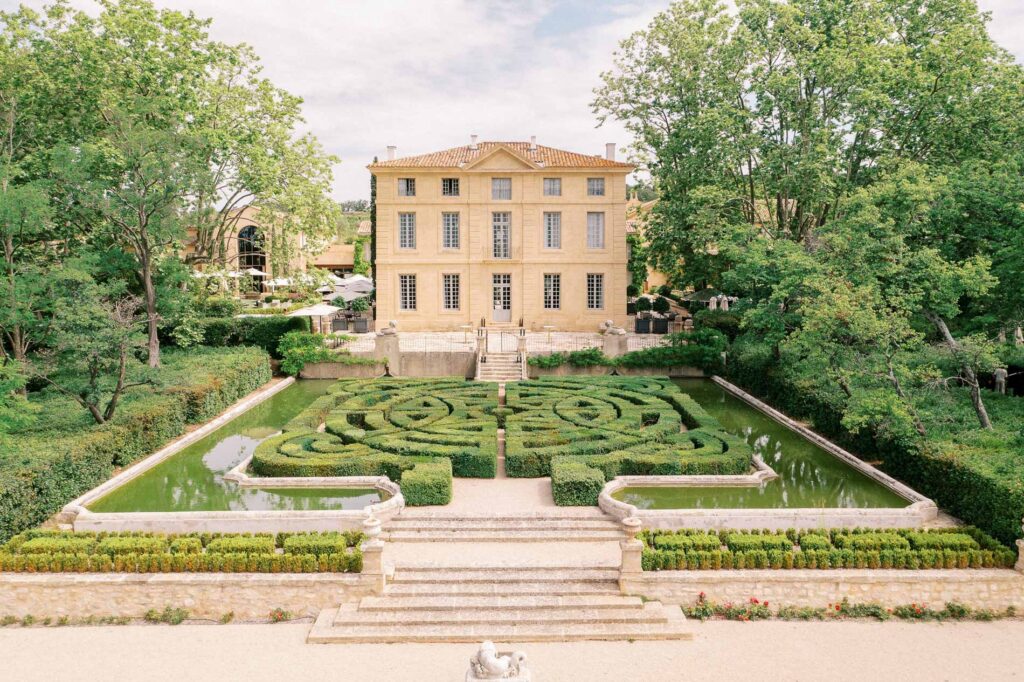 Symmetrical view of Château de la Gaude featuring formal French gardens, sculpted hedges, reflective water features, and an elegant Provençal château surrounded by lush trees.