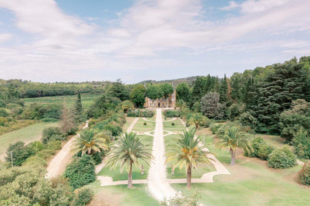Wide aerial landscape shot of Château de Robernier, in the South of France, surrounded by rolling countryside, palm-lined pathways, and lush greenery.