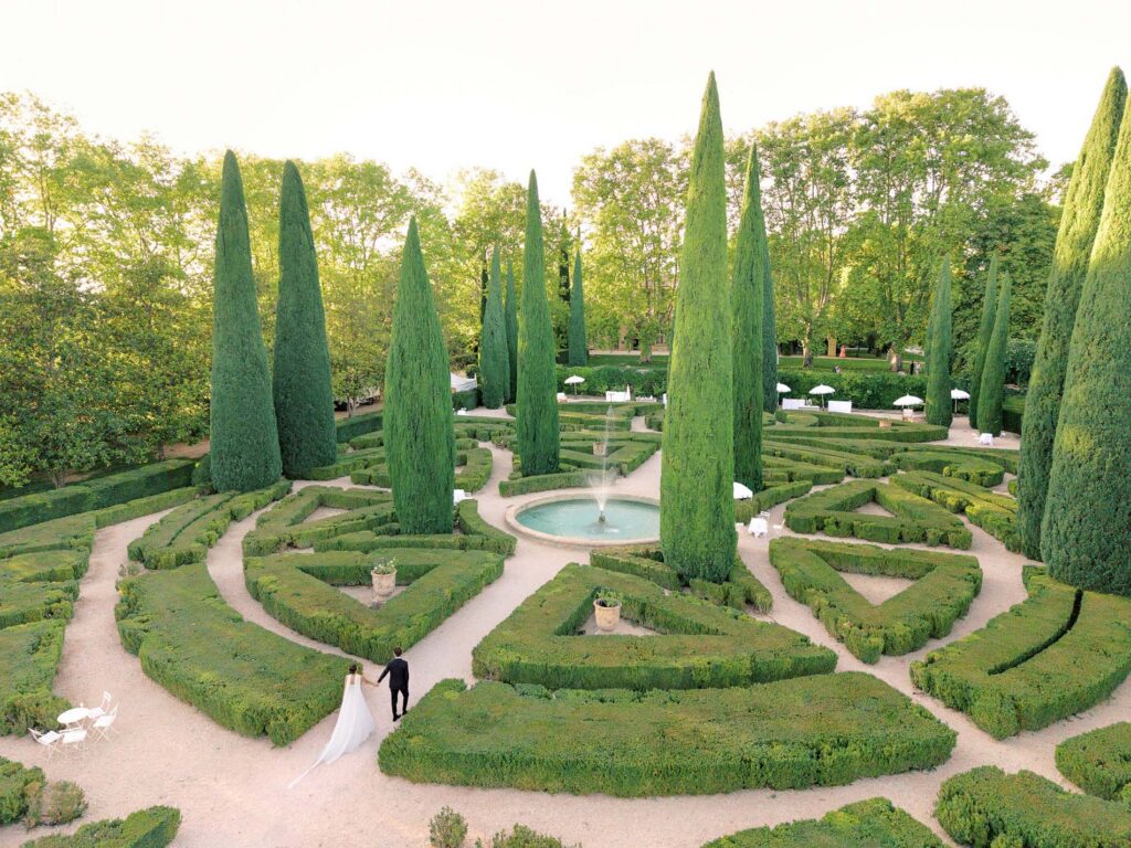 Aerial view of a bride and groom walking through the formal French gardens at Château de Sannes, featuring symmetrical hedge mazes, tall cypress trees, gravel pathways, and a central fountain surrounded by lush greenery in soft golden evening light.