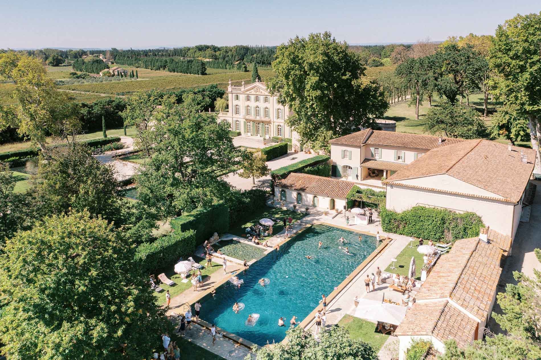 Aerial view of Château de Tourreau in Provence, South of France, during a summer pool party, with guests swimming and lounging beside a long turquoise pool surrounded by manicured gardens, vineyard landscapes, and elegant French estate buildings under warm afternoon sunlight.