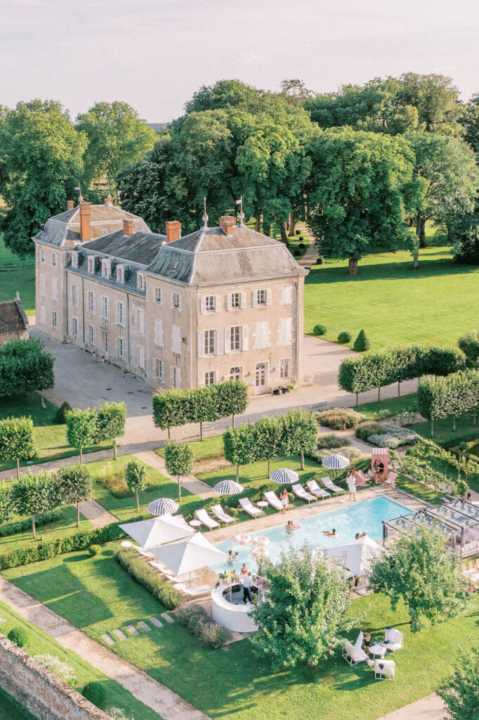 Aerial view of a countryside château during a wedding pool party, with guests relaxing and swimming in a rectangular pool surrounded by sun loungers, umbrellas, and manicured gardens under warm sunlight.