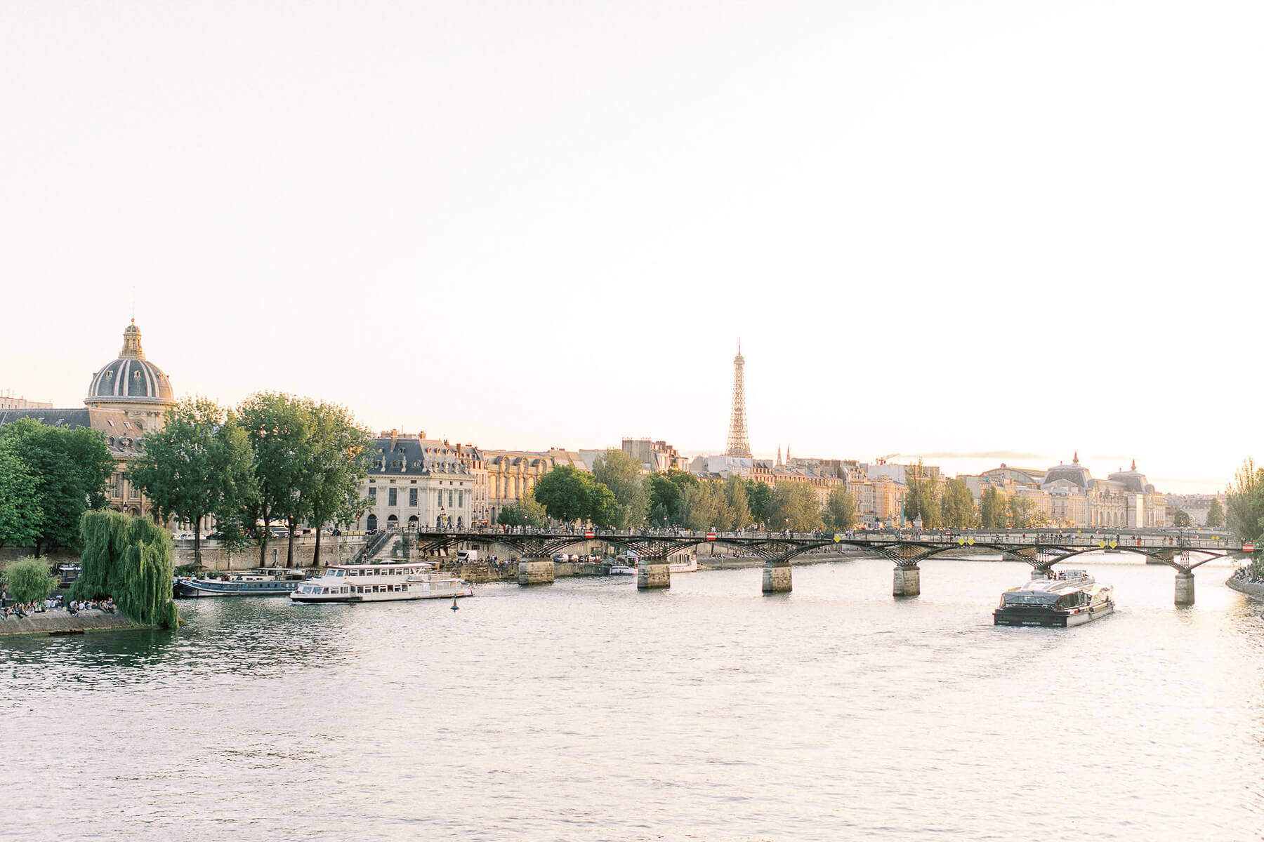 Wide view of the Seine River in Paris at golden hour, with boats cruising past a historic bridge, tree-lined banks, classic buildings, and the Eiffel Tower rising in the distance.