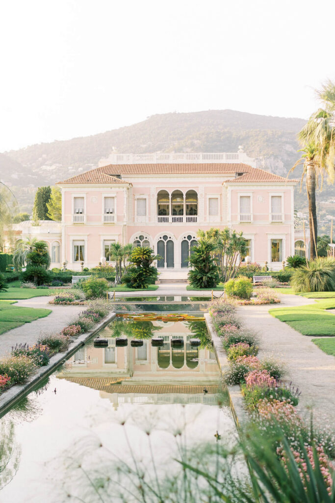 Frontal view of Villa Ephrussi de Rothschild, a pastel pink seaside wedding venue with arched windows and symmetrical architecture, overlooking a long reflecting pool lined with flowers and palm trees, with hills rising in the background.