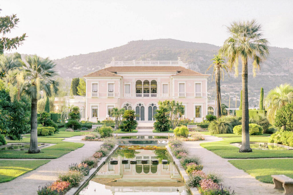 Villa Ephrussi de Rothschild framed by manicured gardens and tall palm trees, with a long reflecting pool leading to the pastel pink villa against sunlit hills in the background. Soft golden-hour light creates a romantic, elegant atmosphere.